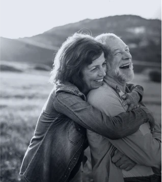 An older couple embracing and laughing in an outdoor setting with hills in the background.