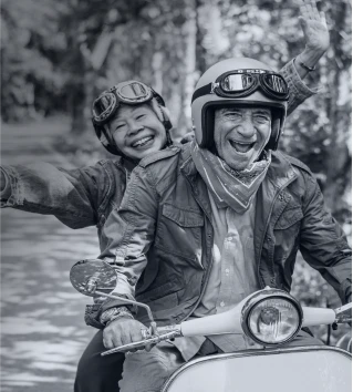 A joyful older couple riding a scooter, both wearing helmets and smiling as they enjoy the ride.
