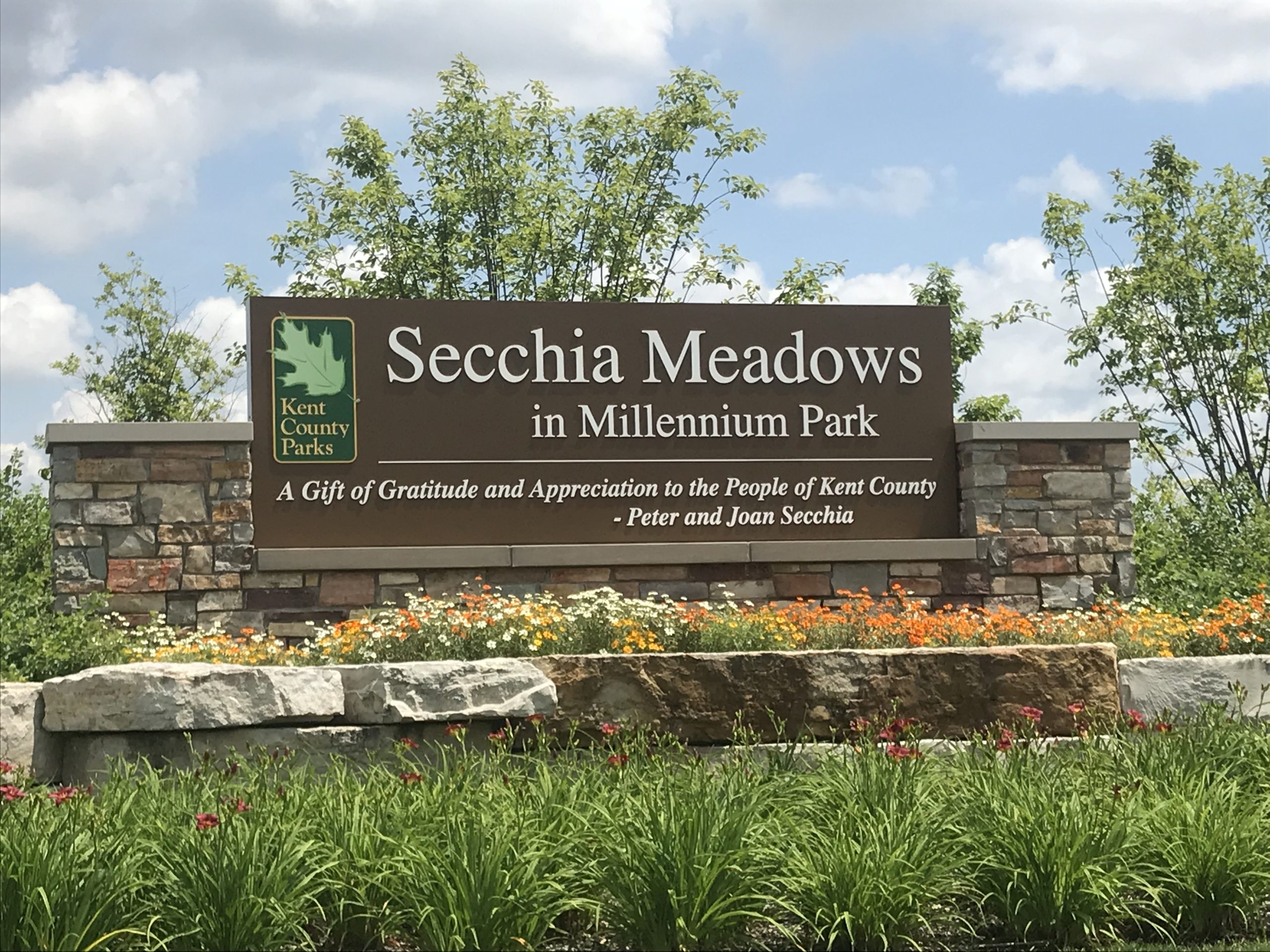 Secchia Meadows sign in Millennium Park, featuring Kent County Parks logo, surrounded by colorful flowers and greenery, representing the venue for the Summer Bash client appreciation event by Jacobs Financial Services.